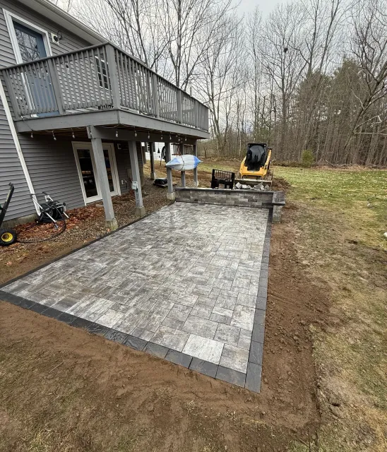 Backyard paver patio beside a gray house with a deck and a compact excavator nearby.