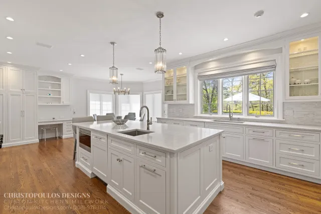 Bright, modern kitchen with white cabinets, large island with marble countertop, stainless steel sink, and large windows showcasing trees outside.