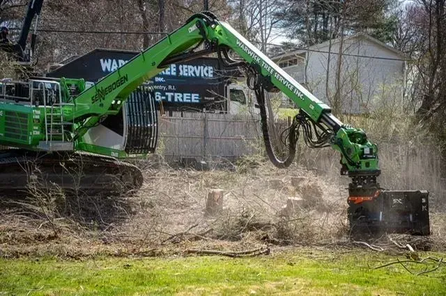 Green tree stump grinder removing stumps in a grassy area, with a white vehicle and a house in the background.