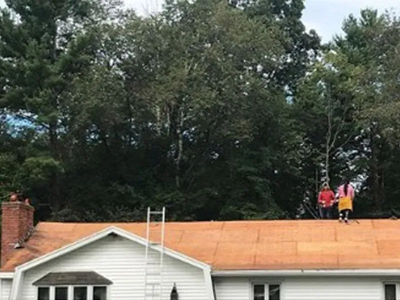 Two workers on a roof with a ladder, re-roofing a white house against a wooded background