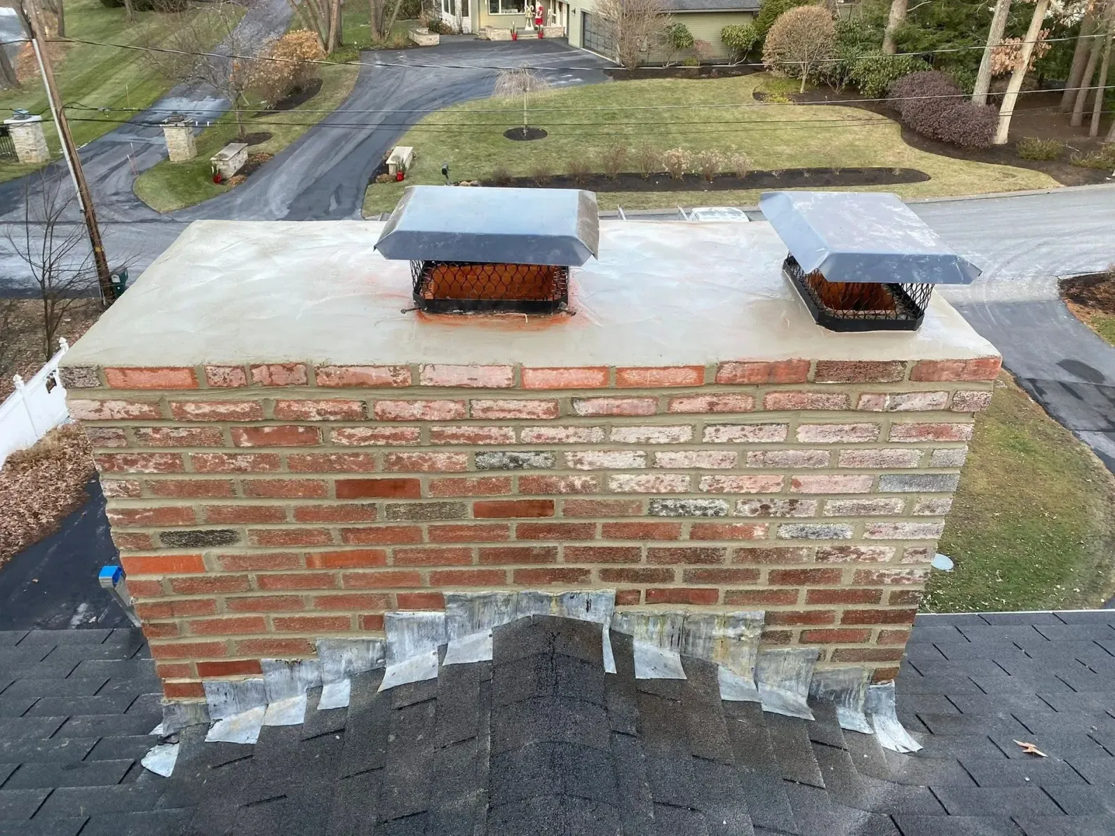 Brick chimney with two metal flue caps on a shingled roof, viewed from above.