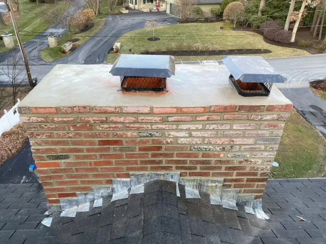 Brick chimney with two metal flue caps on a shingled roof, viewed from above.