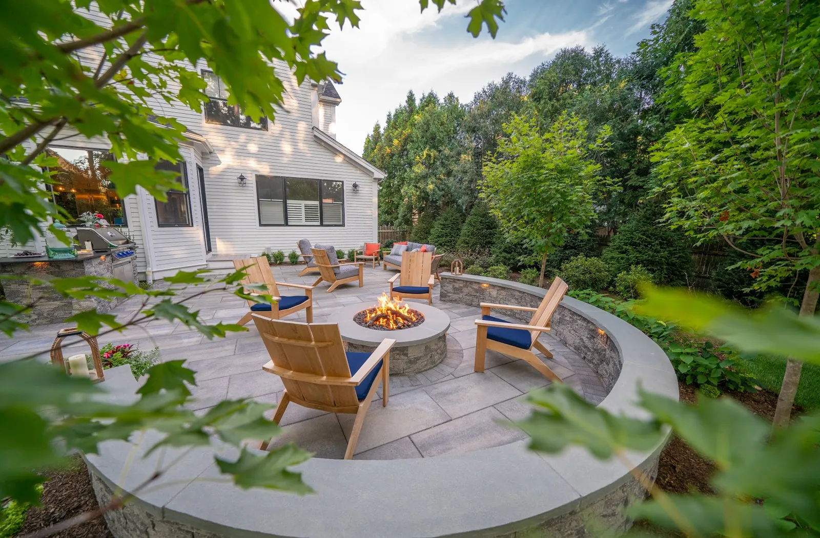 Backyard fire pit patio with curved seating wall and Adirondack chairs surrounded by lush plantings in Needham, Massachusetts by Land Design Associates.