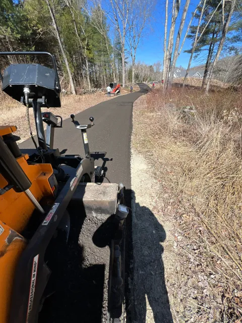 Paved walking path through woods with construction equipment visible on the left and workers in the distance.