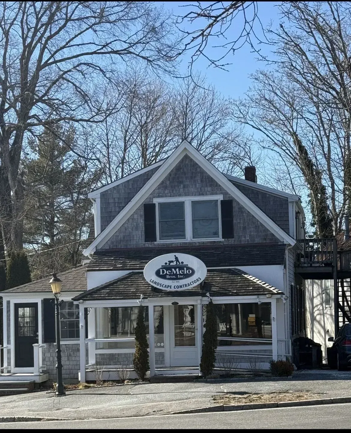 Small gray storefront with a gabled roof and white trim, framed by leafless trees on a sunny day