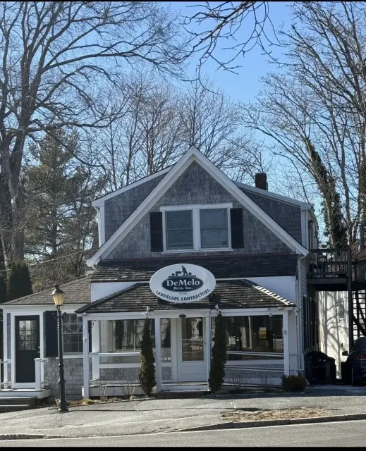 Small gray storefront with a gabled roof and white trim, framed by leafless trees on a sunny day