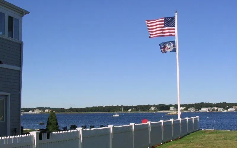 An American flag and a secondary flag wave on a pole beside a white fence overlooking a blue bay on a sunny day.