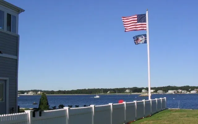 An American flag and a secondary flag wave on a pole beside a white fence overlooking a blue bay on a sunny day.
