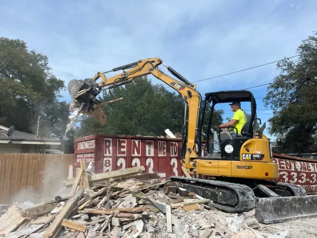 A construction worker using a small excavator to demolish a building with debris and rubble, with a wooden fence and trees in the background.