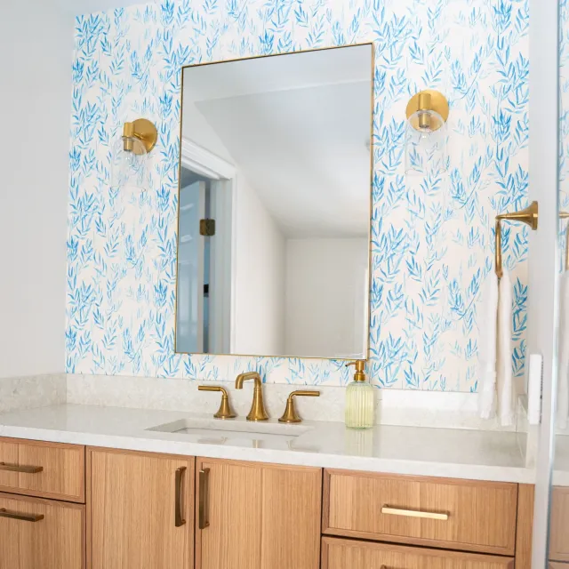 Bathroom with a large mirrored cabinet, gold fixtures, a beige vanity with a white countertop, a soap dispenser, a wall-mounted towel holder with a white towel, and light blue patterned wallpaper.