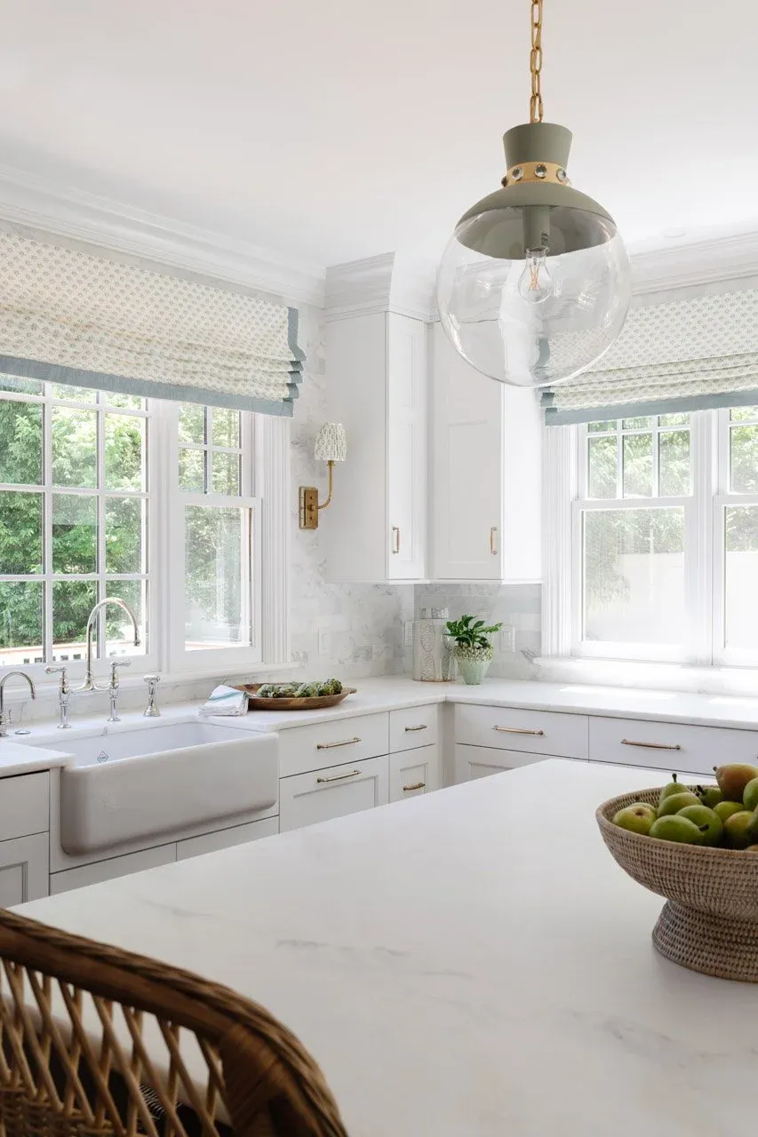 Bright white kitchen with large windows, white cabinetry, and a white marble countertop. A hanging glass pendant light is above the island, and there is a bowl of green apples on the counter.
