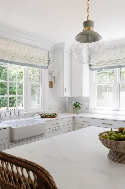 Bright white kitchen with large windows, white cabinetry, and a white marble countertop. A hanging glass pendant light is above the island, and there is a bowl of green apples on the counter.