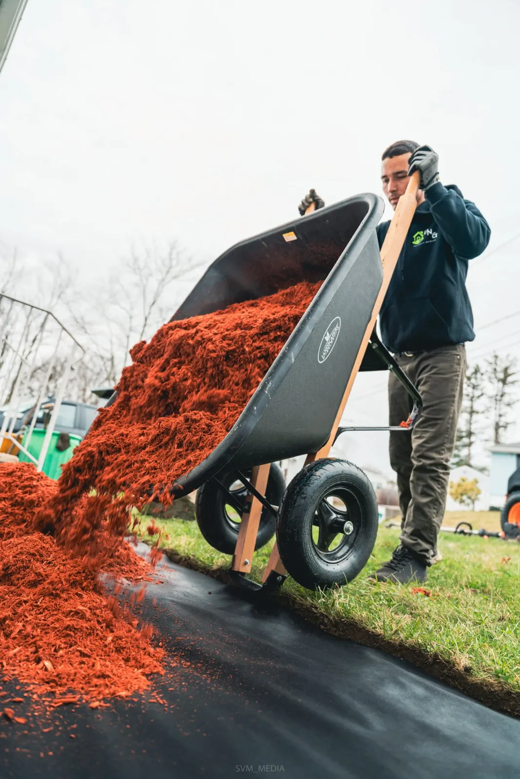 A person using a wheelbarrow to transport reddish mulch on a cloudy day in a yard.