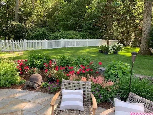 Patio chairs overlooking a lush garden with pink flowers, green lawn, and white fence in sunlight