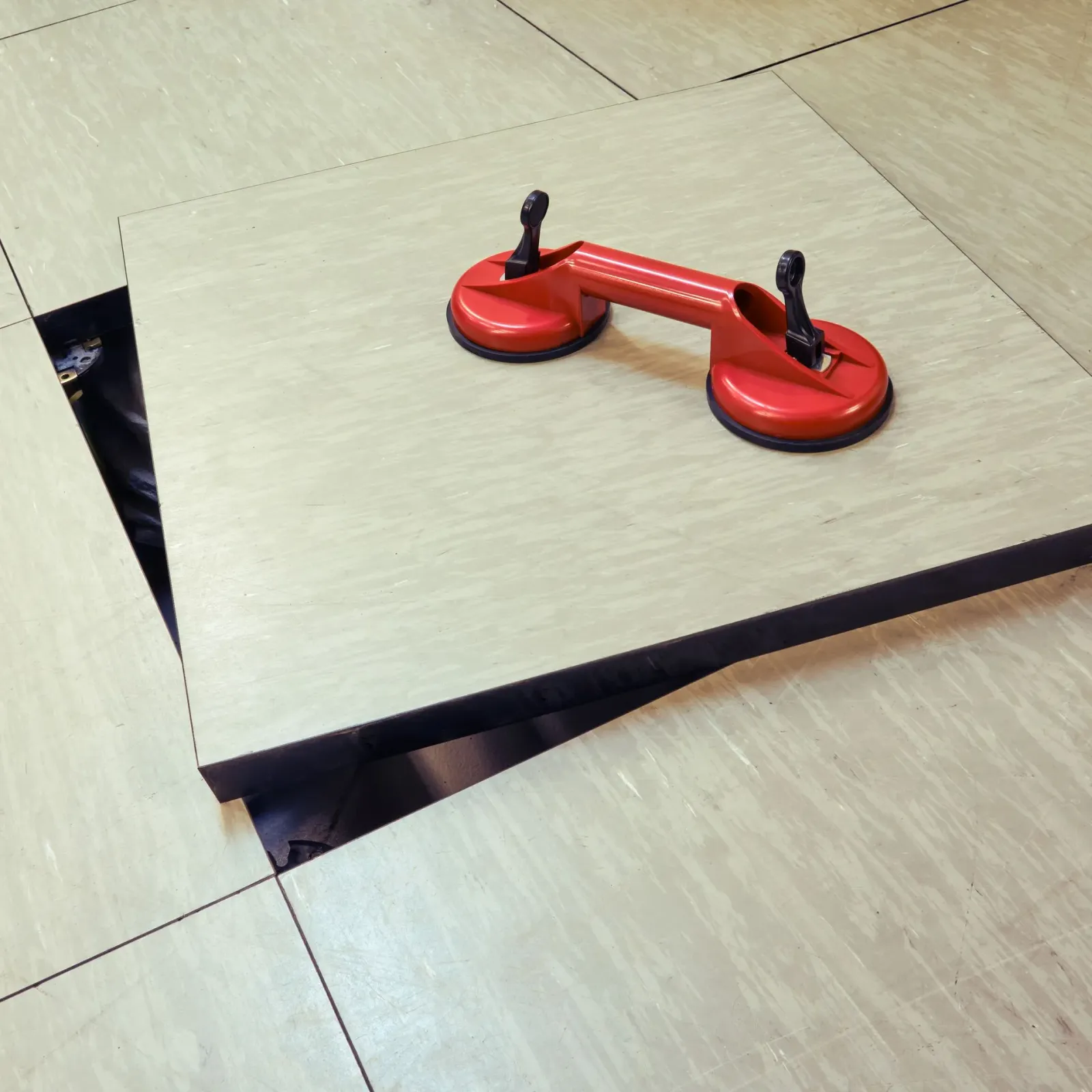 Red suction cup lifter on a light beige floor tile, partially lifted to reveal a dark space below.
