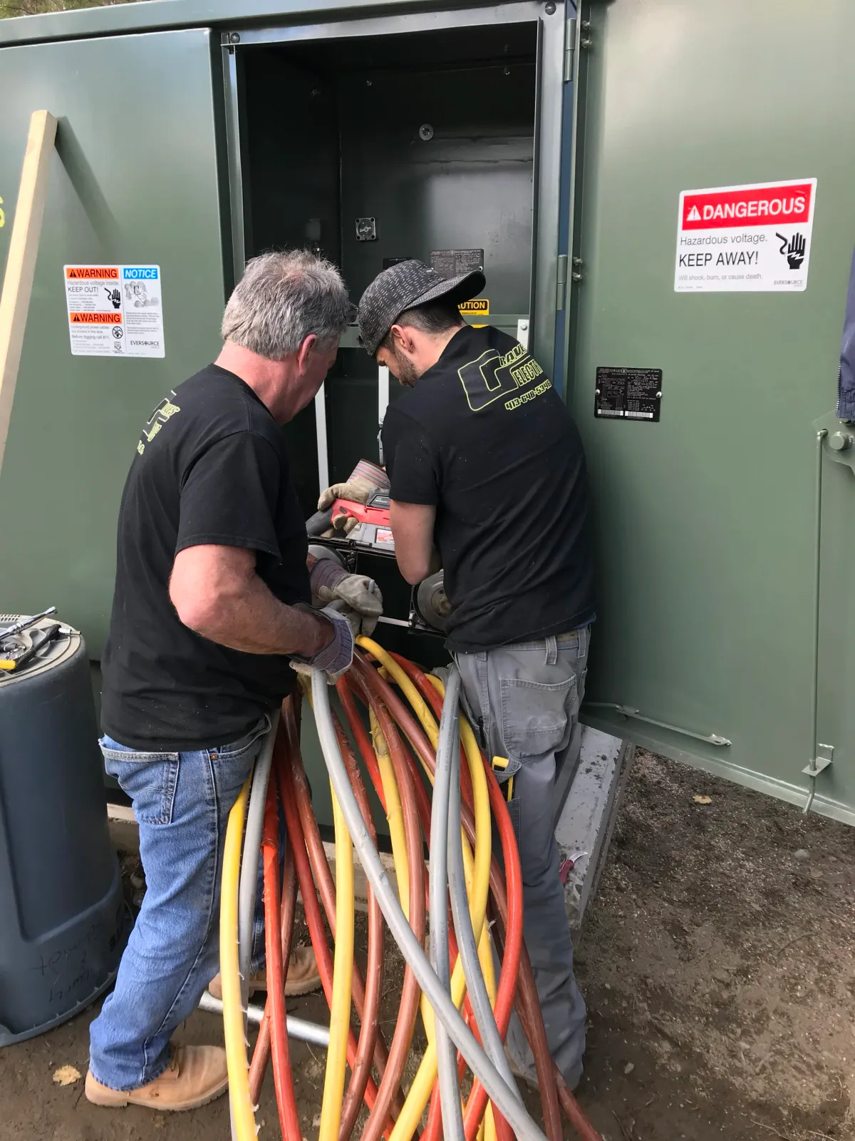 Two workers in black shirts and jeans working on an electrical cabinet outdoors, handling orange, yellow, and gray cables. The cabinet has warning signs about dangerous voltage.