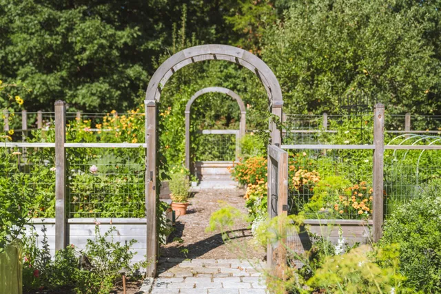 Wooden garden arches lead through open gates along a stone path, flanked by raised beds with colorful flowers and greenery