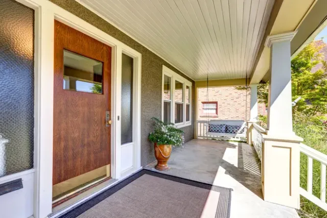 Covered front porch with wooden door, white railings, potted plant, and wicker bench in bright daylight
