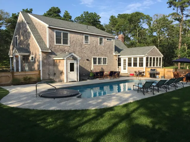 A two-story shingled house with a pool and patio in a wooded backyard.
