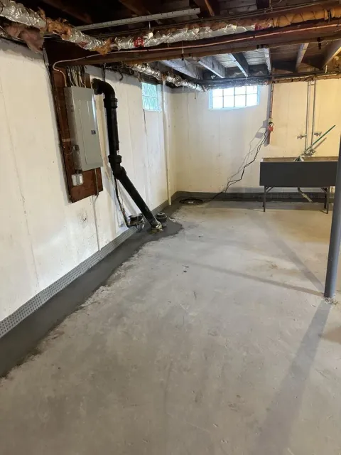 Empty basement with concrete floor, exposed ceiling beams, white walls, and a small window.