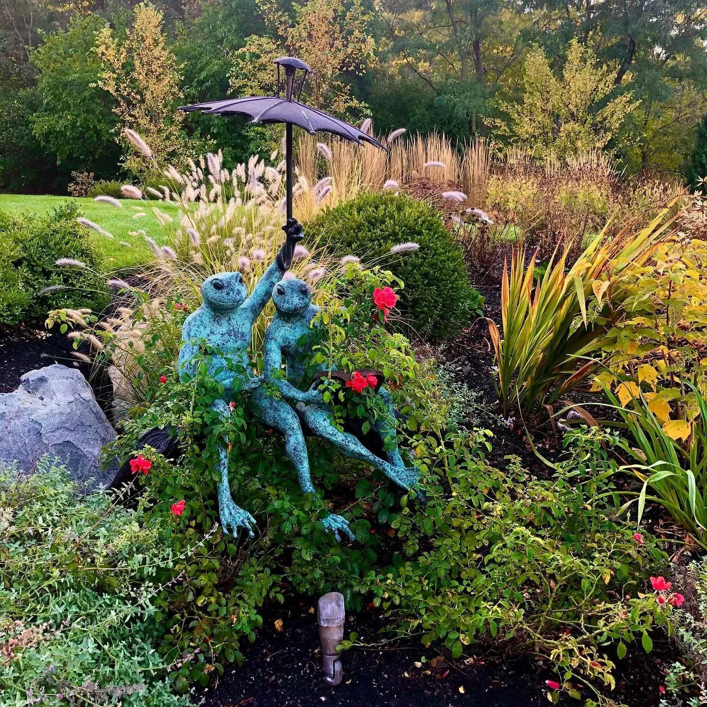Garden scene with a metal sculpture of two frogs sitting on a bench, one frog holding an umbrella over the other, surrounded by various flowers and lush foliage.