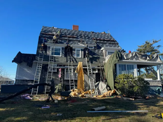 Workers repairing the roof of a two-story house, ladders propped against the siding.