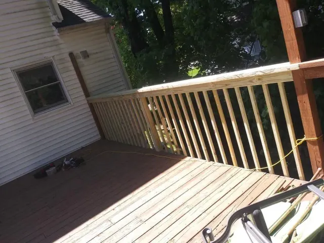 Wooden deck with railing, attached to a house. Sunlight shines on the deck and surrounding trees.