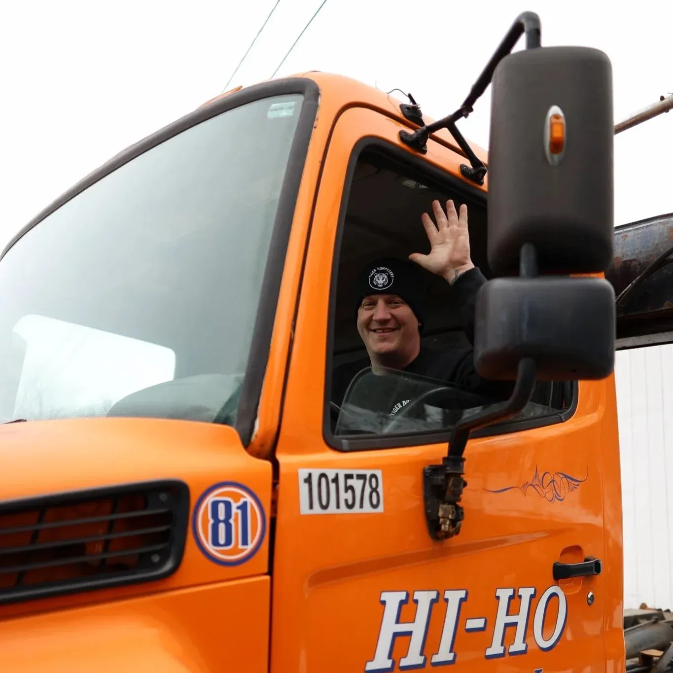 A man in a black beanie and black shirt sitting inside an orange truck, smiling and waving through the window.
