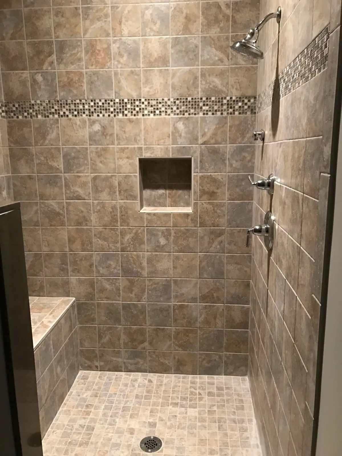 A tiled walk-in shower with a built-in bench, a small square niche, a rain shower head, and two adjustable body spray nozzles.