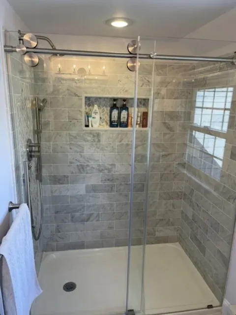 A modern walk-in shower with glass doors, tiled walls, a built-in shelf holding shampoo and body wash bottles, a handheld showerhead, and a window with blinds.