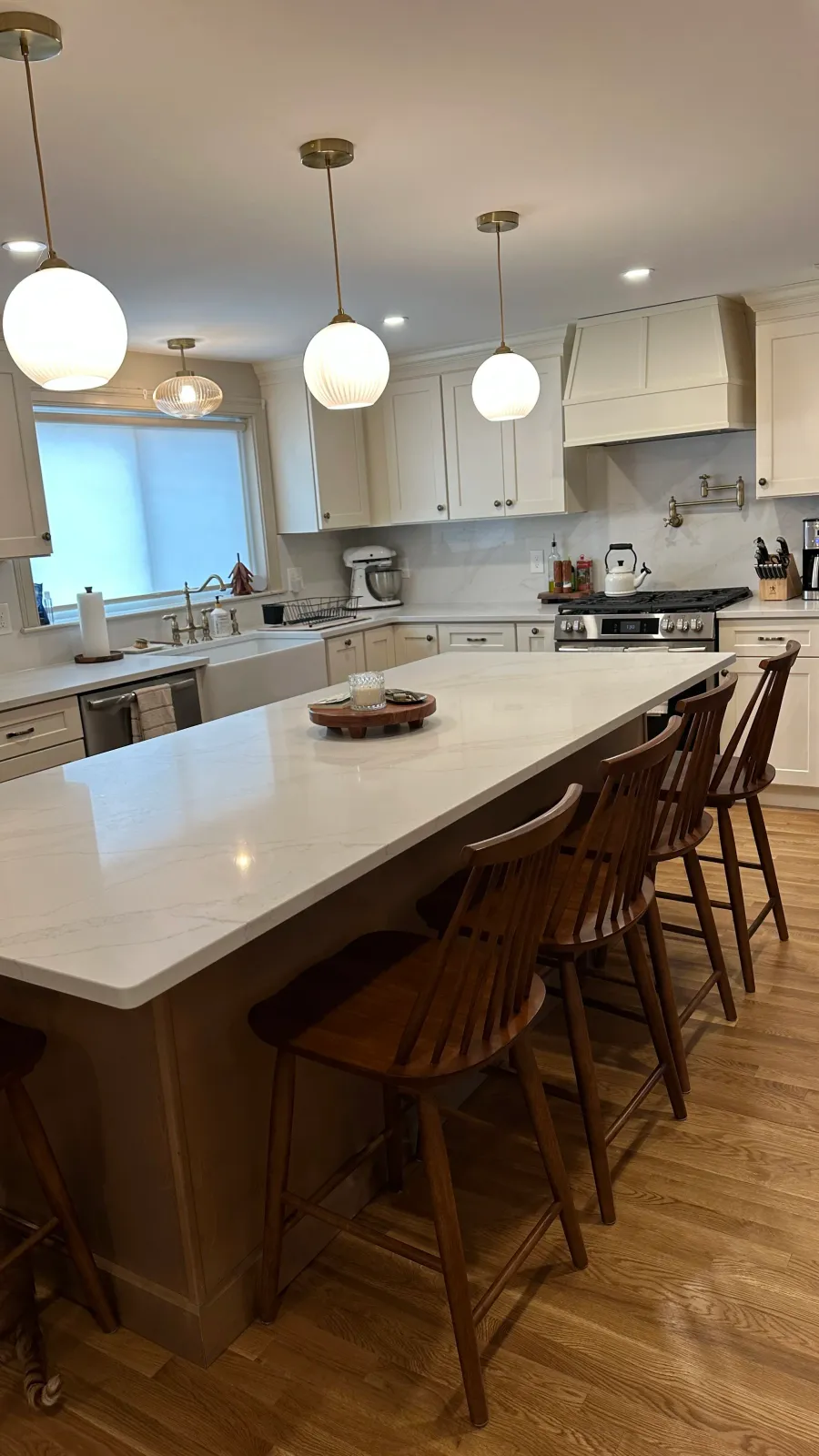 Modern kitchen with white cabinetry, a large marble island with wooden stools, pendant lighting, farmhouse sink, and stainless steel appliances.
