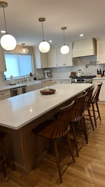 Modern kitchen with white cabinetry, a large marble island with wooden stools, pendant lighting, farmhouse sink, and stainless steel appliances.
