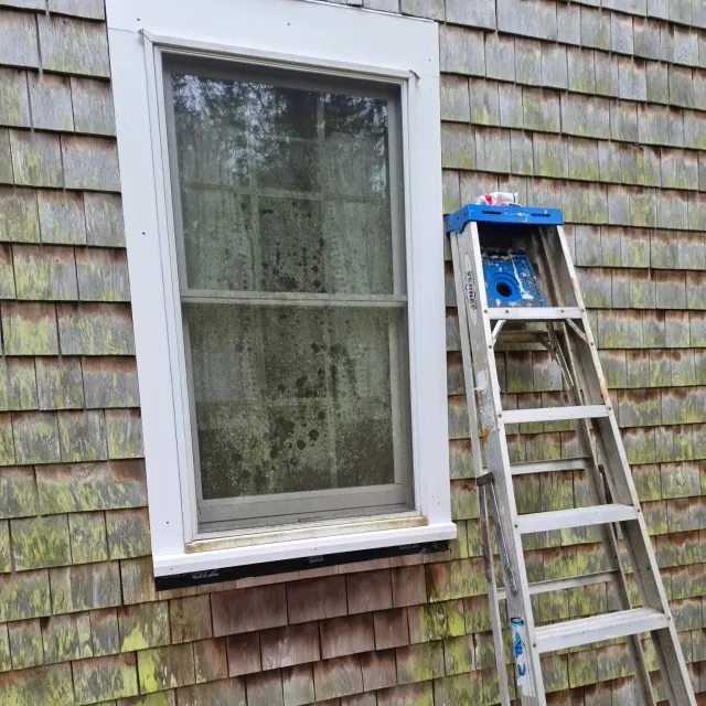 A newly framed window on a house with weathered wooden shingles and a silver extension ladder leaning against the wall beside it.
