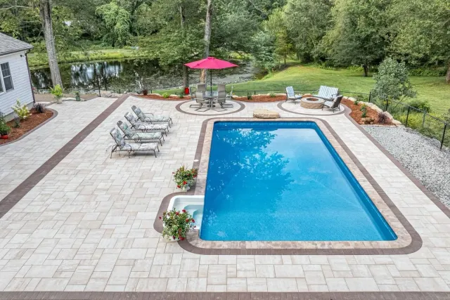 Backyard pool with lounge chairs, red umbrella, and stone patio surrounded by trees