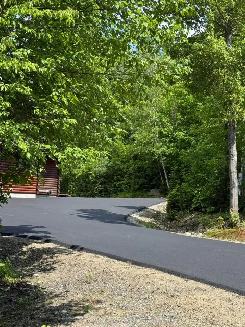 A freshly paved black asphalt driveway curves toward a red wooden building framed by lush green trees.