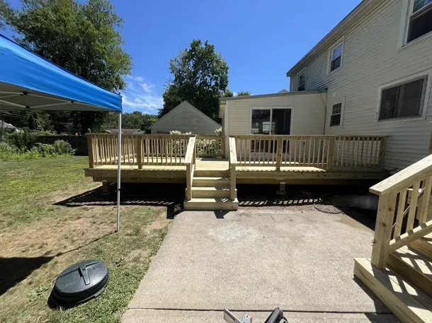 Backyard deck with wooden railings, steps, and a paved patio beside a house under a blue canopy.