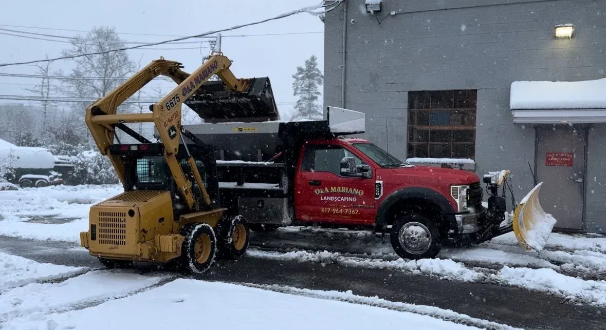 A red landscaping truck with a snow plow attached to the front and a backhoe loader on the bed, clearing snow outside a gray building during snowfall.