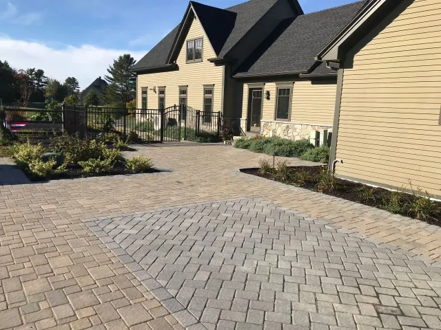 Brick paved driveway leading to a tan house with a dark roof, surrounded by landscaping.