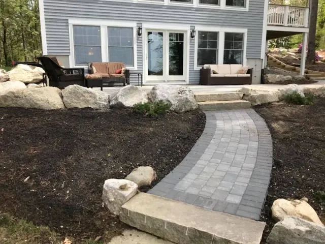 Stone pathway leads to a house with outdoor seating, flanked by large boulders and mulch.