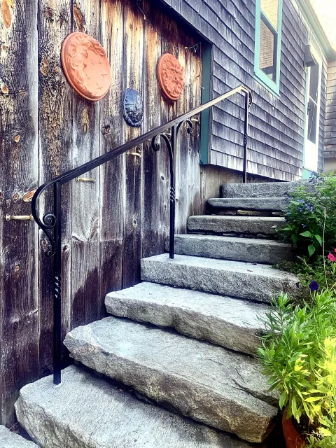 Stone stairs with a black metal handrail leading up to a house with wooden shingles and a window with teal trim. Decorative ceramic plates are mounted on a weathered wooden wall, and there are plants and flowers by the stairs.
