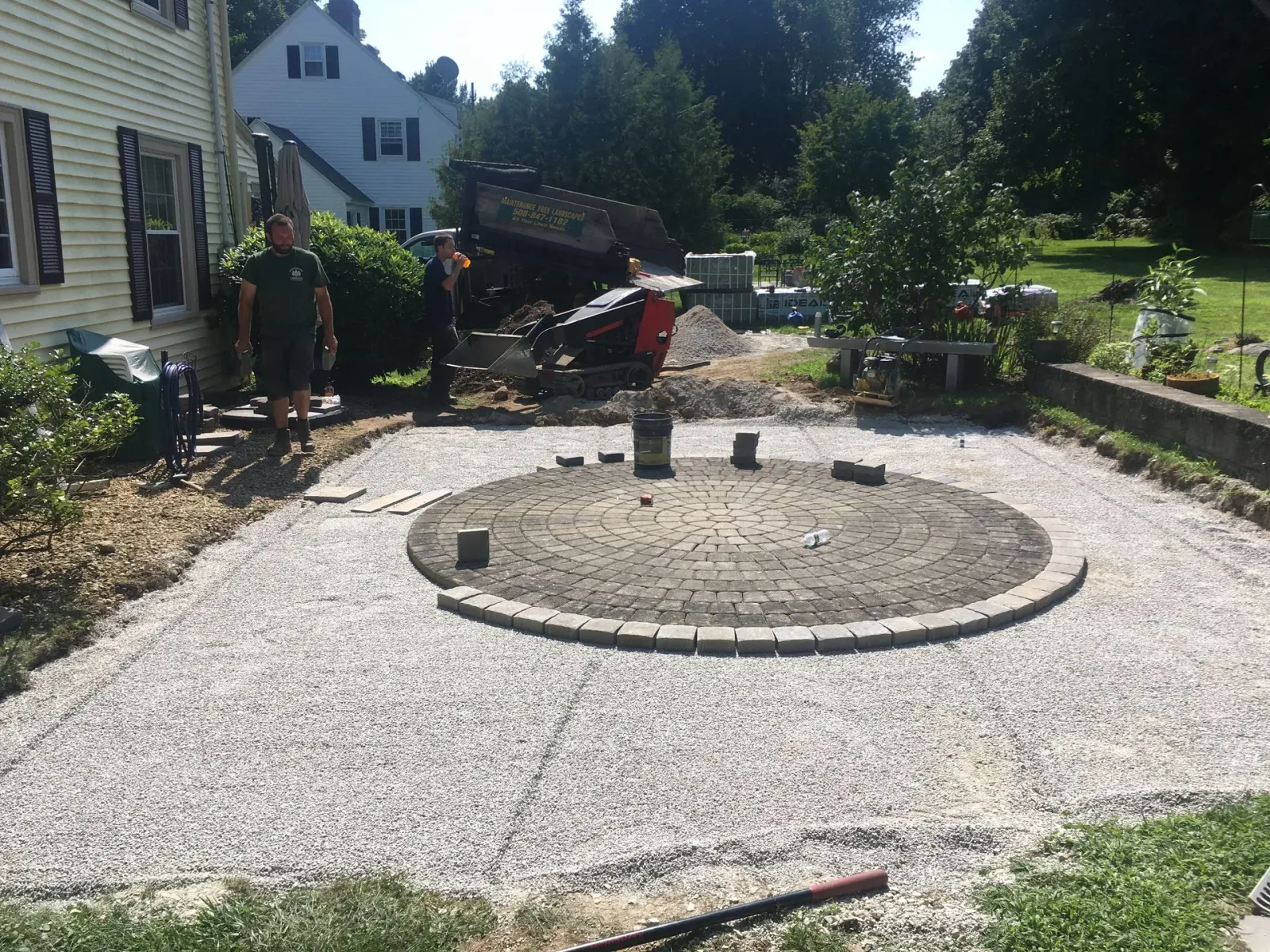 Construction workers stand near a partially completed circular stone patio set in a gravel-filled yard next to a house.