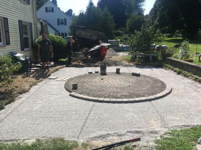 Construction workers stand near a partially completed circular stone patio set in a gravel-filled yard next to a house.