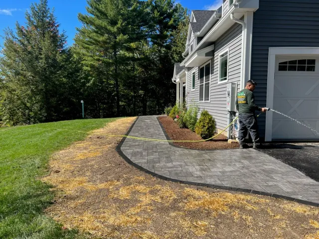 A worker sprays a newly installed paver walkway next to a gray house and a grass lawn under a bright blue sky.