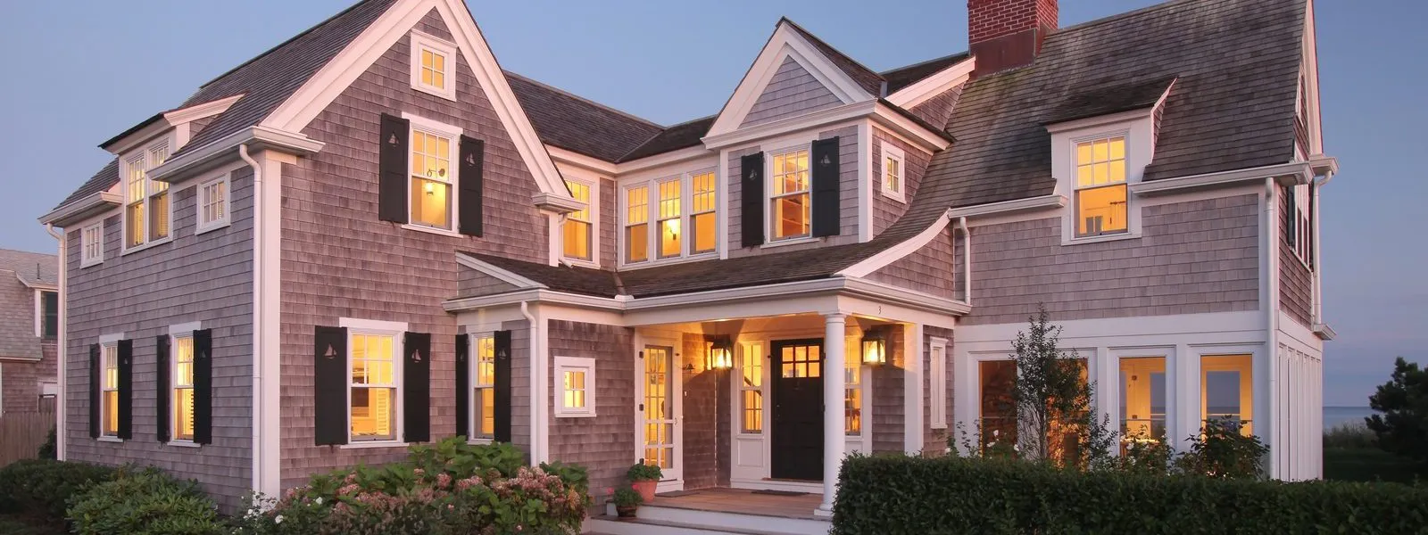 Gray shingle two-story home with white trim and brick chimney at dusk – McPhee Associates of Cape Cod.