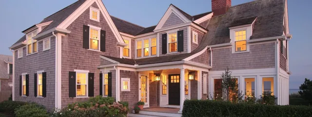 Gray shingle two-story home with white trim and brick chimney at dusk – McPhee Associates of Cape Cod.