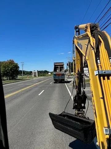 Photo taken from the side window of a construction vehicle showing a road with a truck in front and utility poles on the side under a clear blue sky.