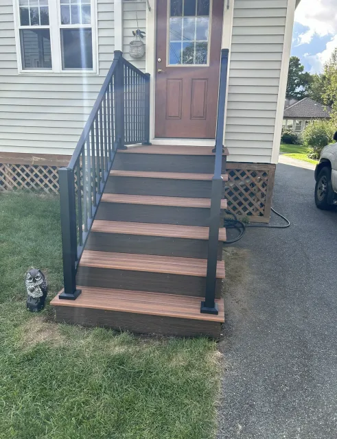 Front porch stairs with black railings leading to a brown door, beside a driveway and lawn.