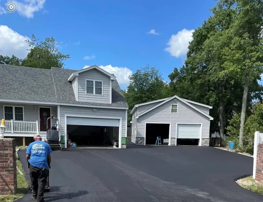 A person in a blue shirt walks toward a house and a detached garage featuring a newly paved, smooth black driveway.