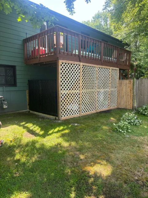 Deck with brown railing over a lattice enclosure, attached to a greenhouse. Lawn in the foreground.