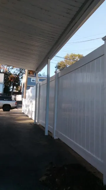 White vinyl fence alongside a covered driveway. Sunlight illuminates the scene, sky in the background.